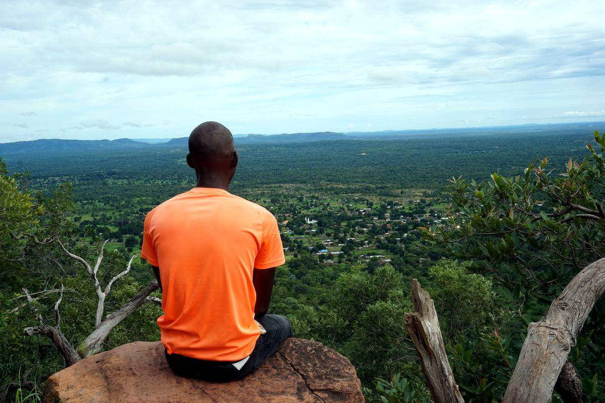 Bouba sentado de espaldas sobre una roca, mirando el valle verde y el pueblo desde un mirador